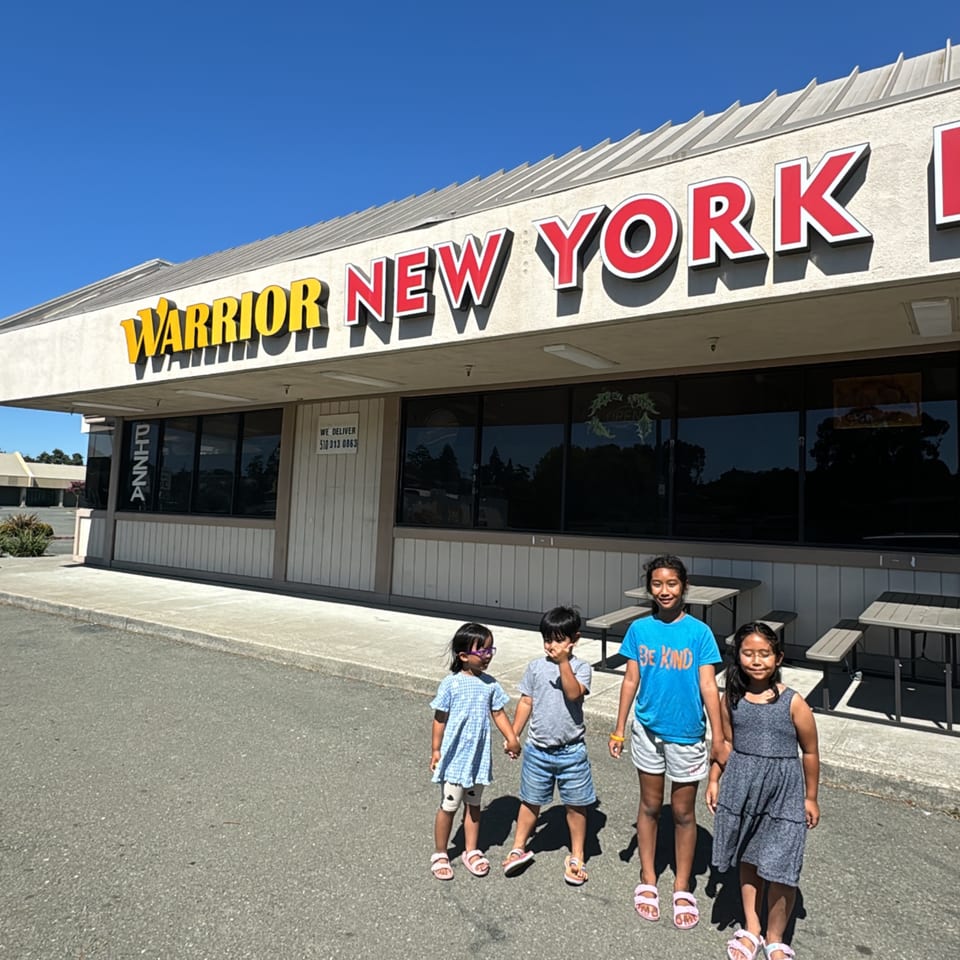 Four children stand in front of the Warrior New York Pizza restaurant on a sunny day.