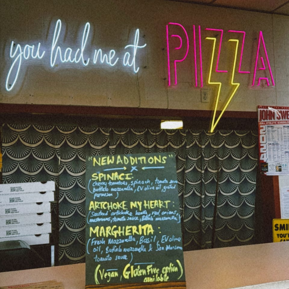 Neon sign at Warrior New York Pizza reads "you had me at PIZZA" above a menu board.
