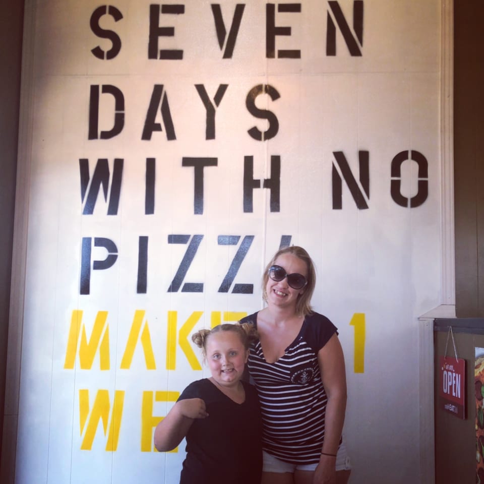 A woman and child stand in front of a wall with a stenciled pizza-themed quote.