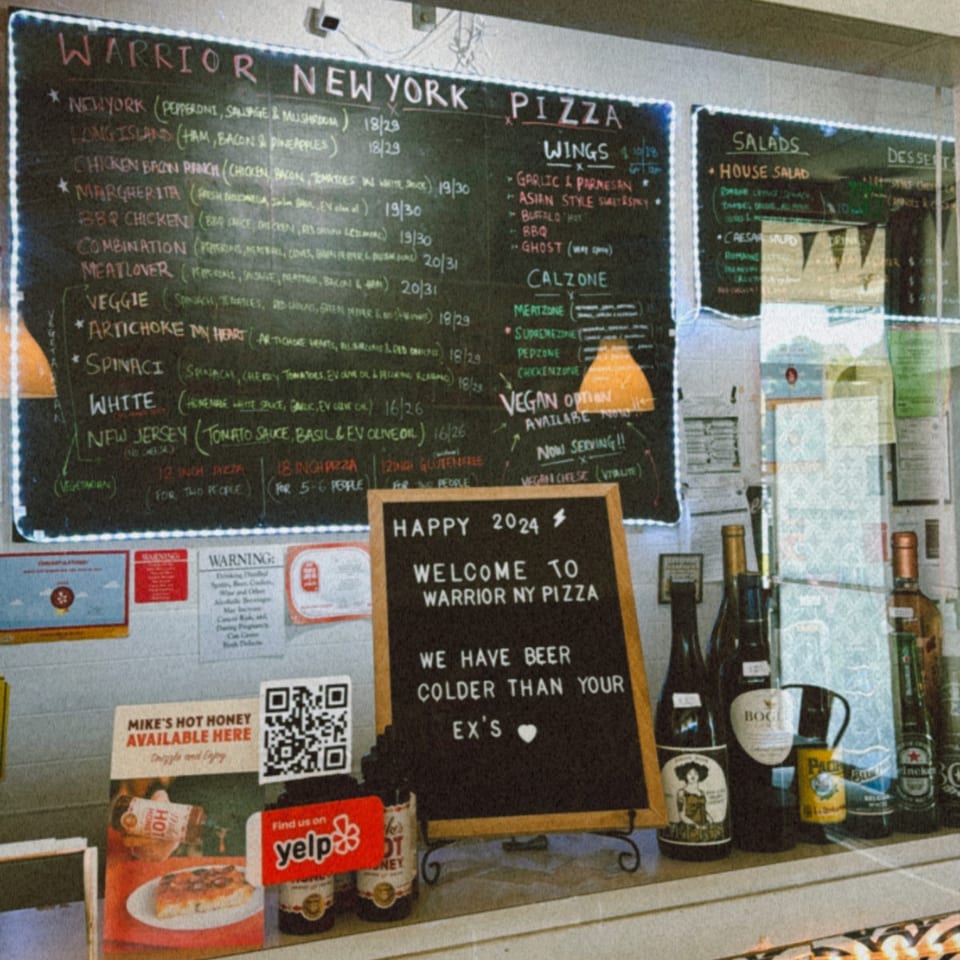 A chalkboard menu for Warrior New York Pizza, displaying pizza, wings, calzones, and salads.
