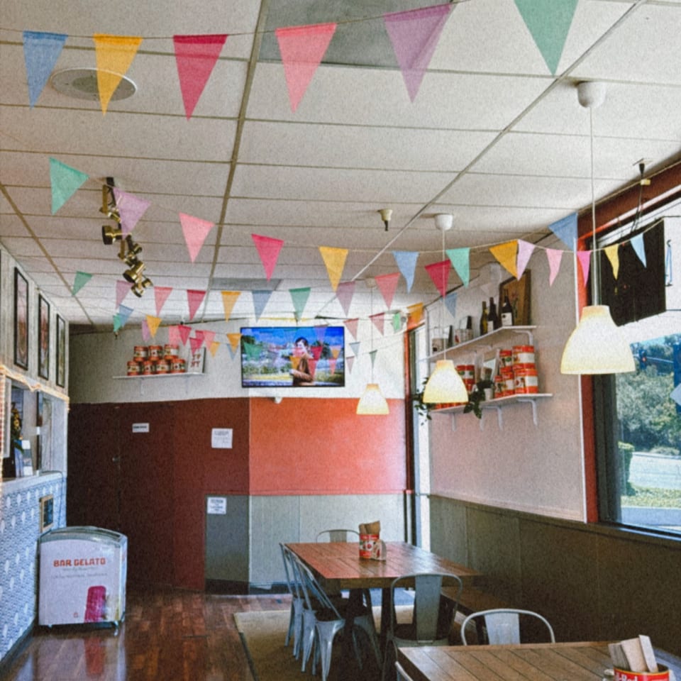 Interior of Warrior New York Pizza with colorful bunting, wooden tables, metal chairs, and a gelato freezer.