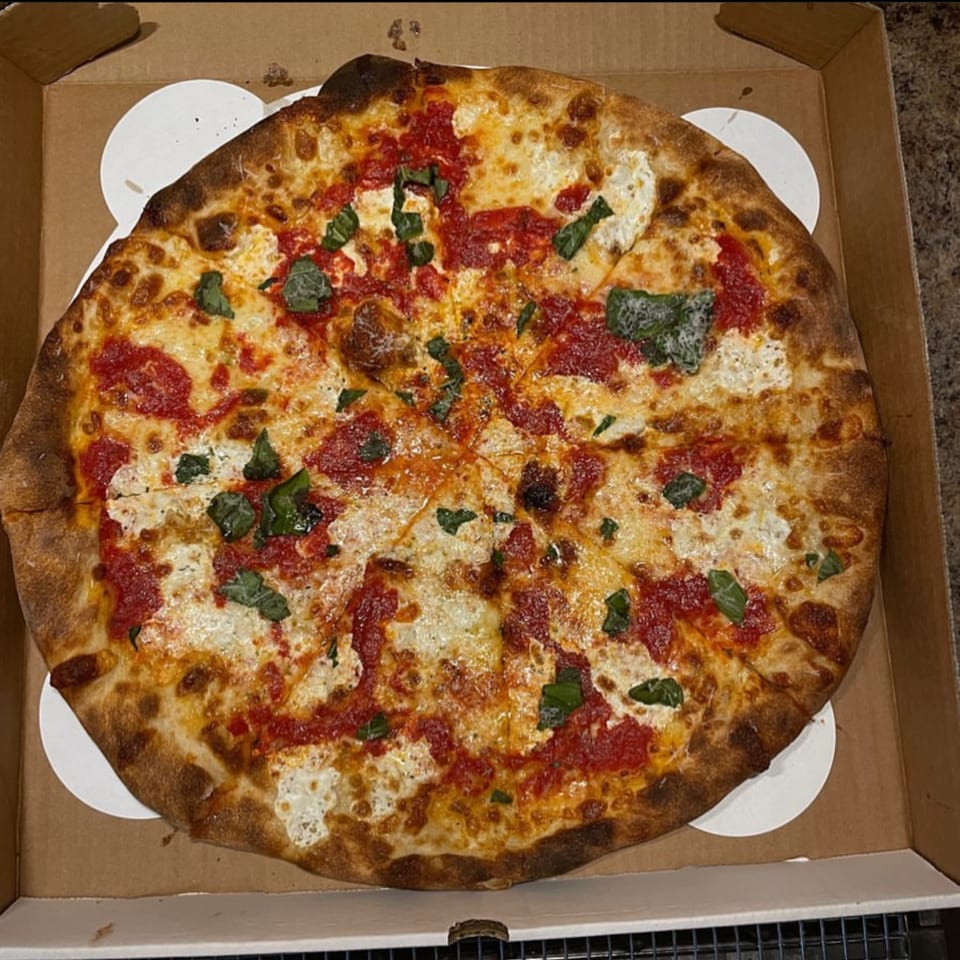 A whole Warrior New York Pizza with red sauce, white cheese, and basil in a cardboard box.